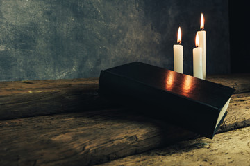 Beautiful closed holy bible and three white burned candles on a old oak wooden table and gray wall background.