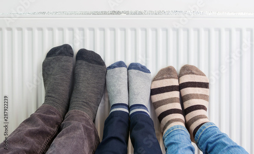 Family wears colorful pair of woolly socks warming cold feet in front of heating radiator in winter time. Electric or gas heater at home. The symbolic image of the heating season. Selective focus.