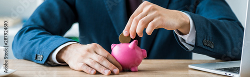 Cuadros en Lienzo panoramic shot of businessman putting metallic coin into piggy bank near laptop