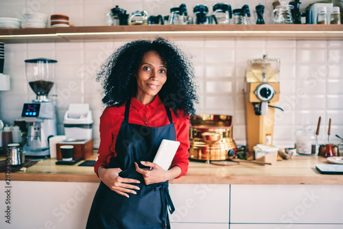 Fotomural Portrait of successful female barista in black apron holding modern touch pad device and smiling at camera standing on working place in own coffee shop
