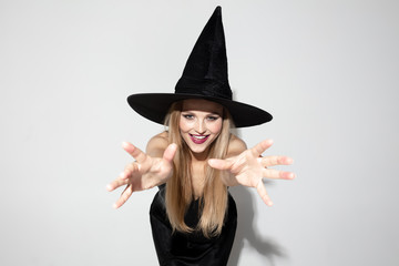 Young blonde woman in black hat and costume on white background. Attractive caucasian female model posing. Halloween, black friday, cyber monday, sales, autumn concept. Copyspace. Looks scary, smiling