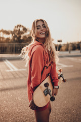 Happy stylish girl with skateboard in red hoodie on city street. Positive fashion young woman outdoors portrait with longboard in sunset evening
