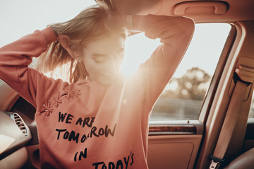 Beauty portrait of pretty girl in car on parking in evening. Stylish urban woman model posing in red hoodie in summer sunset