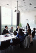 © Maskot - Businesswoman interacting to female colleague sitting with coworkers at conference table during meeting in board room