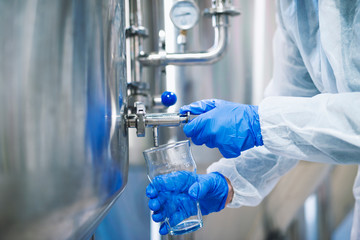 Close up view of hands in protective gloves opening tap valve and filling glass with samples from industrial machine reservoir. Quality control and sampling by technologist. Beverage production.