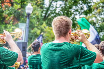 Young clean cut men in sweaty green t shirts play trumpets in parade with bokeh trees and American flag in background - back view and selective focus