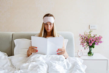 Wall Mural - Beautiful Happy Young Woman reading lifestyle magazine While Lying In Bed After Waking Up In Morning. Portrait Of Smiling Blonde Girl in stylish pajamas in bedroom