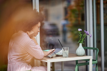 Wall Mural - Grogeous mixed race woman in pink striped dress sitting in cafe and using tablet. On table are cup of coffee, book and lemonade.