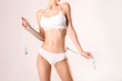 © Evrymmnt - Close up shot of unrecognizable fit woman in lingerie measuring her waist with a tape isolated on white background. Torso of slim attractive female with flat belly in white underwear. Copy space.