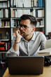 © Dusko - Young nice looking student or writer sitting in college library next to the window and working on his laptop. He is serious and tired. Wall with books in the background.