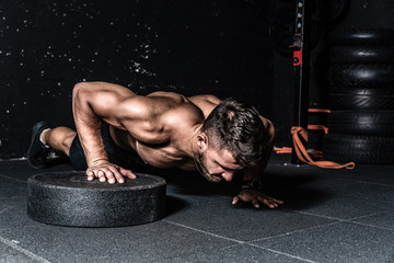 Young strong sweaty focused fit muscular man with big muscles doing push ups with one hand on the barbell weight plate for training hard core workout in the gym real people selective focus