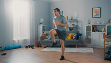 Strong Athletic Fit Man In T-shirt And Shorts Is Energetically Jogging In Place At Home In His Spacious And Bright Living Room With Minimalistic Interior.