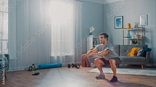 Fotomural Muscular Athletic Fit Man in T-shirt and Shorts is Doing Squat Exercises at Home in His Spacious and Bright Living Room with Minimalistic Interior
