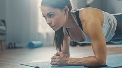 Strong Beautiful Fitness Girl in Athletic Workout Clothes is Doing a Plank Exercise While Using a Stopwatch on Her Phone. She is Training at Home in Her Living Room with Cozy Interior.