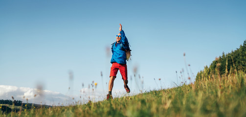 Wall Mural - backpacker traveler emotionally jumping over green grass mountain meadow with backpack with wide ope