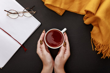 Wall Mural - cropped view of woman holding tea in mug near glasses, scarf and blank notebook on black background