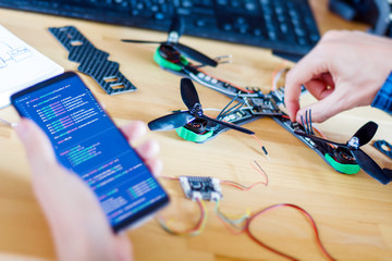 Closeup programmer hand is holding smartphone with program code software on screen for controlling FPV drone. Man developer is building constructing quadcopter from kit with microcontrollers.