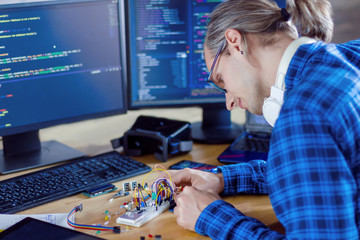 Developer is connecting breadboard to microcontroller for creating prototype of electronic device. Chips, resistors, light-emitting diodes on desktop of hardware engineer. Program code on background.