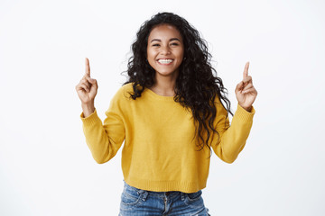 Cheerful optimsitic young female student with curly dark hairstyle, yellow sweater, smiling and laughing happily, pointing fingers up, showing friends link to site or copy space, white background