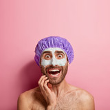 Fototapeta  - Worried young man applies white mud mask on face, feels refreshment and rejuvenation, wears bathcap, stands shirtless against pink background, copy space above. Face care, softness and purity