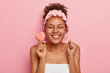 Young overjoyed dark skinned female model clenches fists with happiness, holds small soft heart shaped sponge, closes eyes from pleasure, ready to take bath, wrapped in towel wears headband for shower