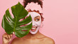 Cropped shot of happy young woman applies facial mud mask on face for removing wrinkless or fine lines, takes bath at bathroom, wears protective rosy headband, holds big plant leaf covers eyes with it