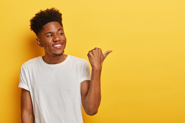 Wall Mural - Friendly cheerful dark skinned hipster man discusses interesting copy space and points thumb away on right side, shows some place or direction, has curly trendy haircut, wears casual white t shirt