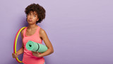 Sad unhappy dark skinned woman exercises with hula hoop, holds fitness mat rolled up under arm, has no wish for workout, wears pink vest, isolated over purple studio wall with copy space aside