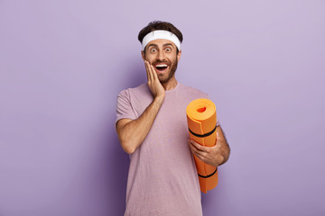 Positive Caucasian man stands with rolled up karemat, touches cheek, wears headband and t shirt, stands against purple background has fitness workout regularly, waits for coach, motivated for training