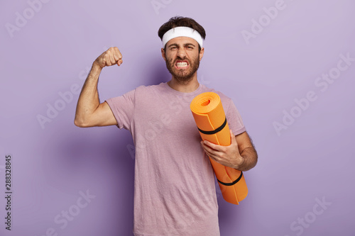Fotografía Motivated powerful man stands with fitness mat, enjoys yoga as sport and hobby, raises arm and shows muscle, clenches teeth, wears headband, violet t shirt