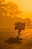 Fototapeta Zwierzęta - A backlit Mozambican cyclist with dramatic shadow, carrying a heavy load of coal to market along a tarmac road at sunrise. Nampula Town, Mozambique