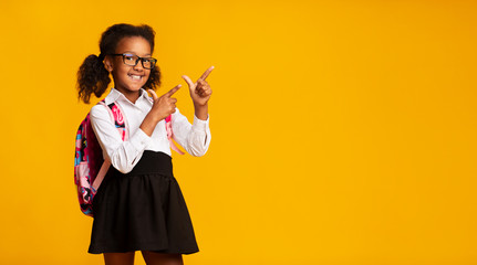 Cheerful Black Schoolgirl Pointing Fingers At Copyspace, Yellow Background