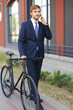 © ty - Portrait of a smiling young business man dressed in suit talking on mobile while standing with bicycle outdoors.