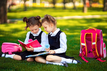 Two Little School Girls With Pink Backpack Sitting On Grass After Lessons And Read Book Or Study Lessons, Thinking Ideas, Education And Learning Concept.