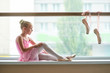 Lovely young ballerina on blurred background. Ballet girl sitting on window-sill. Pair of pointe shoes hanging on a training barre.