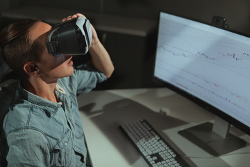 Young man using virtual reality goggles, sitting in front of his compputer late at night. Male freelancer resting from working late, using vr headset, copy space
