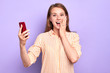 Front view of suprised funny young woman smiling broadly, holding red cell phone, cant believe in her success, happy and emotional, dressed in stylish shirt, standing at light purple wall.