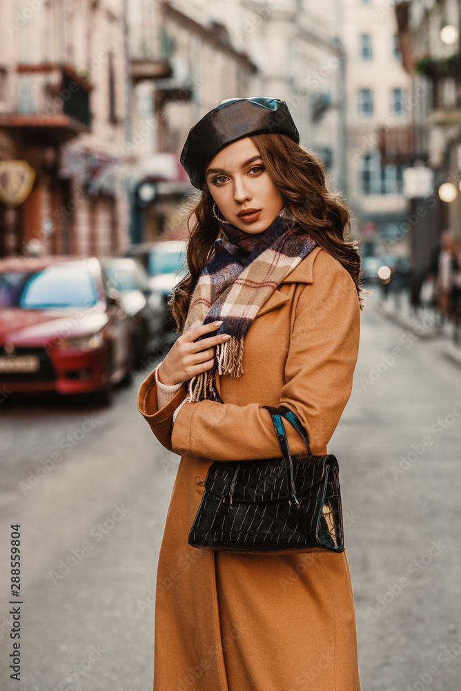 Outdoor autumn portrait of young elegant fashionable lady wearing trendy beret, camel color coat, plaid scarf, holding textured reptile faux leather baguette handbag, posing in street of European city - obrazy, fototapety, plakaty 
