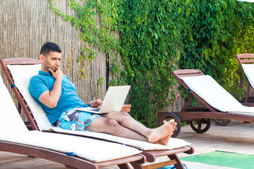 Wall Mural - Portrait of clever handsome bearded young adult freelancer man in blue t-shirt and shorts lying on cozy deck chair with laptop on poolside, working online and finding solution in his head. Lifestyle