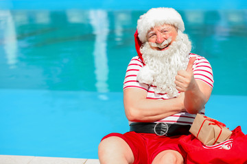 Wall Mural - Santa Claus with gifts showing thumb-up near swimming pool at resort