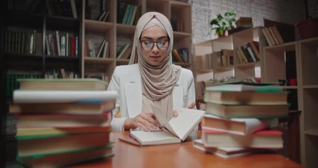 Wall Mural - Female muslim student writes down notes in her notebook, sitting at desk covered with books and looking at camera - modern islam, student lifestyle concept close up 4k portrait