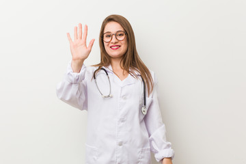 Young doctor woman against a white wall smiling cheerful showing number five with fingers.