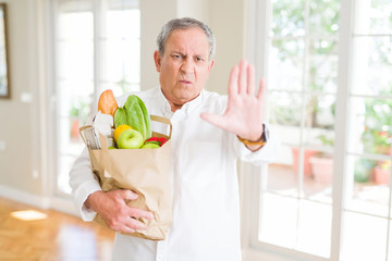 Wall Mural - Handsome senior man holding a paper bag of fresh groceries from the supermarket with open hand doing stop sign with serious and confident expression, defense gesture