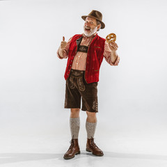 Wall Mural - Portrait of Oktoberfest senior man in hat, wearing the traditional Bavarian clothes. Male full-length shot at studio on white background. The celebration, holidays, festival concept. Eating puff.