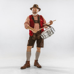 Wall Mural - Portrait of Oktoberfest senior man in hat, wearing the traditional Bavarian clothes. Male full-length shot at studio on white background. The celebration, holidays, festival concept. Playing drums.