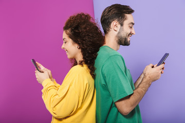 Wall Mural - Portrait of pleased caucasian couple in colorful clothing smiling and holding cellphones standing back to back