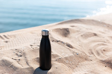 Matte Black Steel Thermo Bottle For Water In Sand Of The Beach On Background Of Sea. Empty Blank For Mockup.