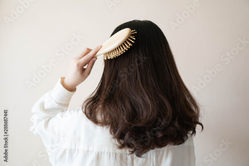 Back view of young beautiful woman in white shirt with long black curly hair combing her hair in the morning. Hair care concept.
