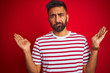 © Krakenimages.com - Young indian man wearing striped t-shirt standing over isolated red background clueless and confused expression with arms and hands raised. Doubt concept.