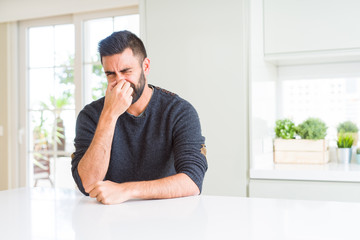 Canvas Print - Handsome hispanic man wearing casual sweater at home smelling something stinky and disgusting, intolerable smell, holding breath with fingers on nose. Bad smells concept.